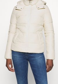 Giacca puffer beige chiaro con chiusura a zip, cappuccio regolabile e polsini elasticizzati. Presenta una trapuntatura orizzontale e tasche laterali.