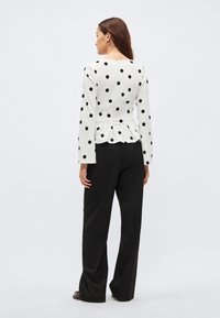 Hvit bluse med svarte polkadotter, lange ermer og peplum i midjen; kombinert med løse svarte bukser. Enkel design og glatt stofftekstur.