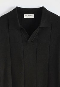 Polo noir en tissu texturé, avec un design en col en V et un col ribbé, étiquette de marque visible au niveau du col. Taille M.