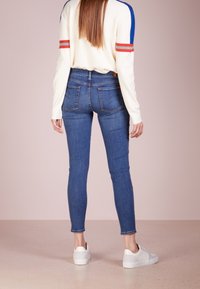 Mujer de pie de espaldas, vistiendo zapatillas blancas, jeans ajustados azules y una camiseta blanca de manga larga con rayas rojas y azules en las mangas.