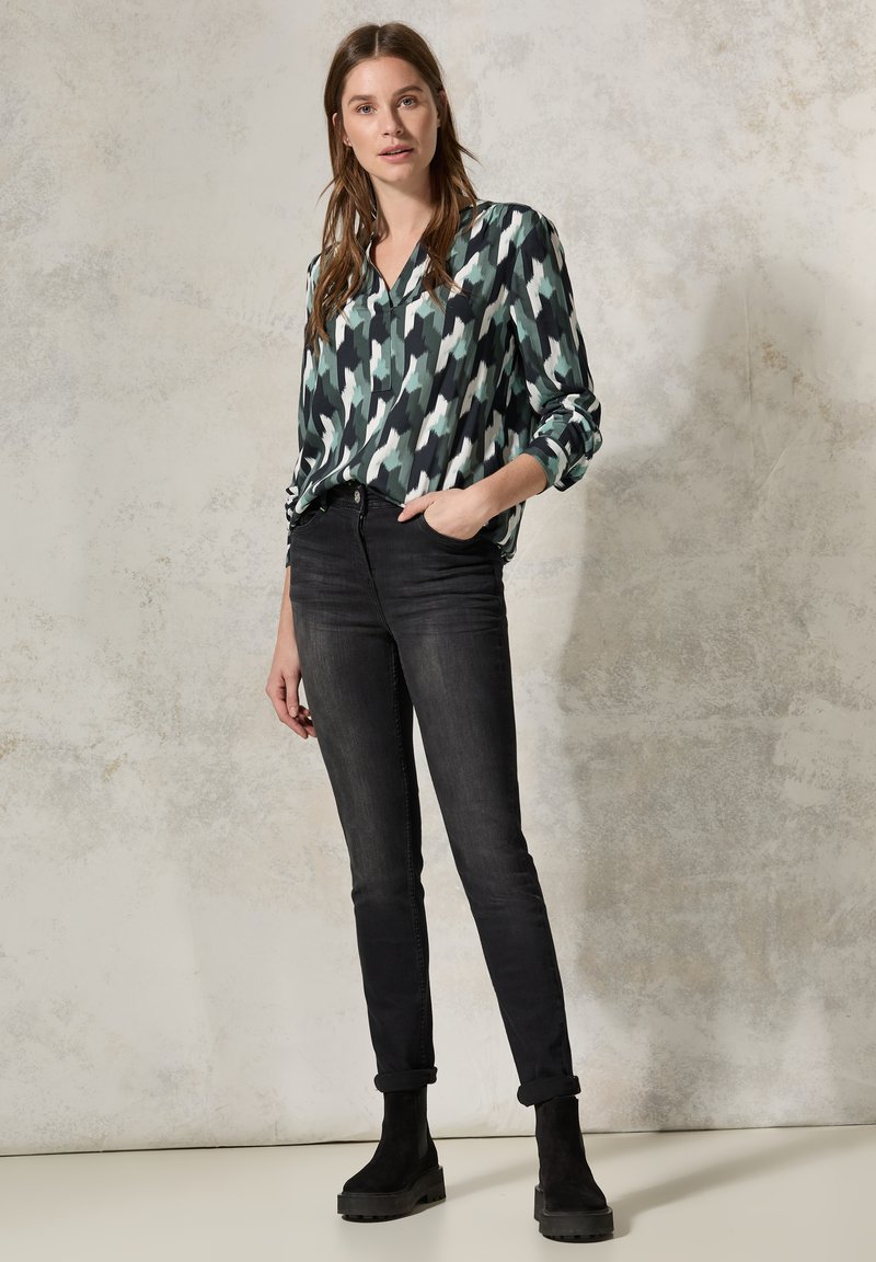 Blusa a maniche lunghe con una fantasia in nero, verde e bianco; abbinata a jeans neri aderenti e stivaletti neri alla caviglia. Design minimalista e tessuto leggero.