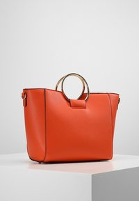 Oranžová kožená taška na rameno se strukturálním tvarem, zdobená výraznou zlatou kruhovou rukojetí a minimalistickým designem. Hladký povrch, žádné viditelné vzory.