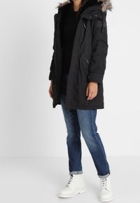 Sort parka jakke med hætte trimmet med pels, frontlynlås, sidelommer og justerbare manchetter, parret med blå jeans og hvide ankelstøvler.