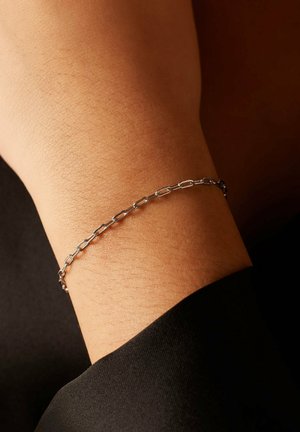 Zilveren kettingarmband met verlengde schakels, gedragen om een gebruinde pols. Glad oppervlak tegen een donkere stof achtergrond, benadrukt het minimalistische ontwerp.