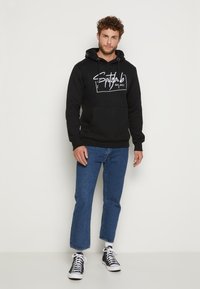Schwarzer Hoodie mit einer Vorderentasche, weißem grafischem Text, verstellbarem Kapuzenzug und gerippten Bündchen. Kombiniert mit blauen Jeans und schwarzen Sneakern.
