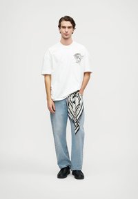 Camiseta blanca con estampado gráfico, jeans rectos de color azul claro y zapatos negros; bufanda de rayas en blanco y negro atada a la cintura.