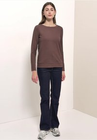 Braunes, geripptes Langarmshirt kombiniert mit dunkelblauen, ausgestellten Jeans. Das Model trägt silberne Sportschuhe und steht vor einem neutralen Hintergrund.