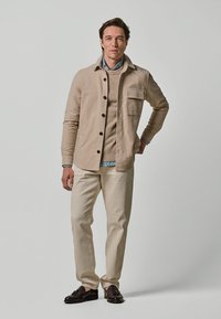 Beige knap-jakke med en frontlomme, båret over en beige sweater og en lyseblå skjorte, parret med cremede bukser og mørke sko.