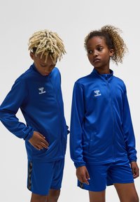 Blaue Sportjacken mit durchgehendem Reißverschluss, kombiniert mit passenden Shorts. Die Jacken verfügen über Reißverschlusstaschen und ein Logo. Strukturierter Stoff für Komfort.