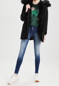 Černá parka s umělou kožešinou na kapuci, zelený svetr s vzorem a modré gradient skinny džíny. Model má na sobě bílé tenisky. Jednoduchý, ležérní outfit.