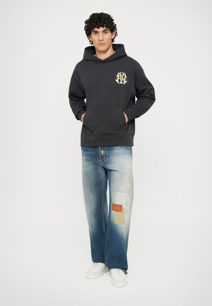 Sudadera gris oscuro con bolsillo delantero y logo en contraste. Jeans ajustados azules claros con parches decorativos. Zapatillas blancas y fondo natural.