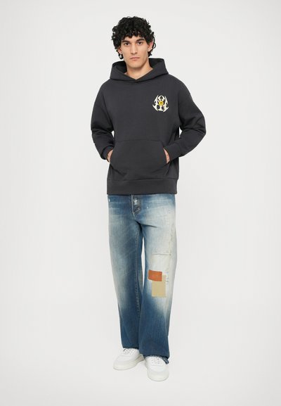 Dark gray hoodie with front pocket and contrasting logo. Light blue distressed jeans with patch accents. White sneakers and natural backdrop.