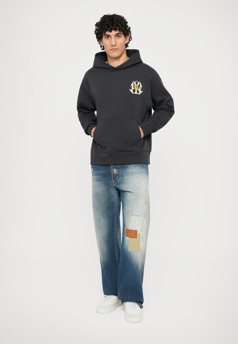 Felpa grigia scura con tasca frontale e logo a contrasto. Jeans azzurri chiari e strappati con dettagli a toppa. Sneakers bianche e sfondo naturale.