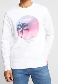 Sweat-shirt blanc avec un graphique circulaire représentant un palmier et des couleurs pastel, marqué "Levi's California" en haut, avec poignets côtelés.