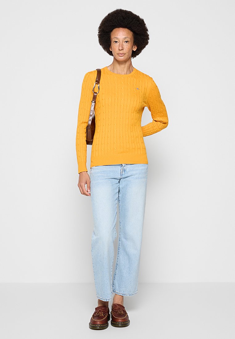 Sweter w kolorze żółtym, o splocie warkoczowym, dopasowany krój, długie rękawy. Jasnoniebieskie jeansy typu straight-leg. Brązowe buty. Brązowa torba na ramię. Minimalne oznakowanie na swetrze.