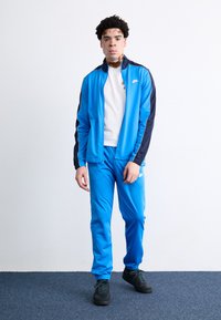 Μπλε φόρμα με φερμουάρ και μαύρες λεπτομέρειες, ταιριαστά παντελόνια και μαύρα αθλητικά παπούτσια. Το υλικό φαίνεται λείο και αθλητικό.