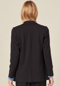 Blazer noir structuré avec un col cranté, des manches longues et une fente dans le dos. Présente des poignets de chemise bleu clair contrastants.