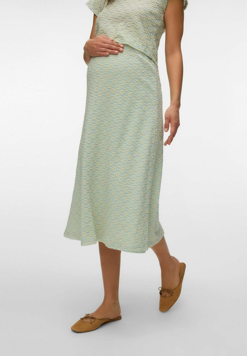 Femme enceinte portant une robe midi à motifs vert clair et des chaussures plates marron clair, posant avec une main sur son ventre, sur fond blanc.