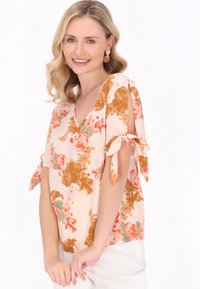 Blusa floreale in morbido tessuto pesca, con scollatura a V, maniche corte con dettagli a fiocco e un motivo di fiori arancioni e rosa.