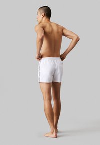 Pantaloncini da bagno bianchi con cintura elastica, taglio fino al ginocchio e strisce laterali. Texture morbida, design minimale e senza dettagli hardware visibili.