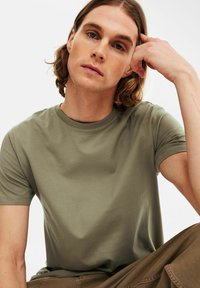 Oliven grøn T-shirt i et blødt, glat stof med rund hals og korte ærmer, båret af en person med bølget hår, siddende.