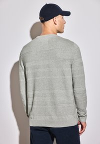 Pull gris avec un motif de rayures texturé, col rond et manches longues. Porté avec une casquette marine, vu de l'arrière.