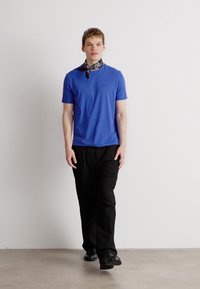 Μοντέλο που φοράει μπλε κοντομάνικο μπλουζάκι και μαύρο παντελόνι. Ένα διακοσμημένο κασκόλ είναι δεμένο γύρω από τον λαιμό. Λείο ύφασμα, άνετη εφαρμογή.