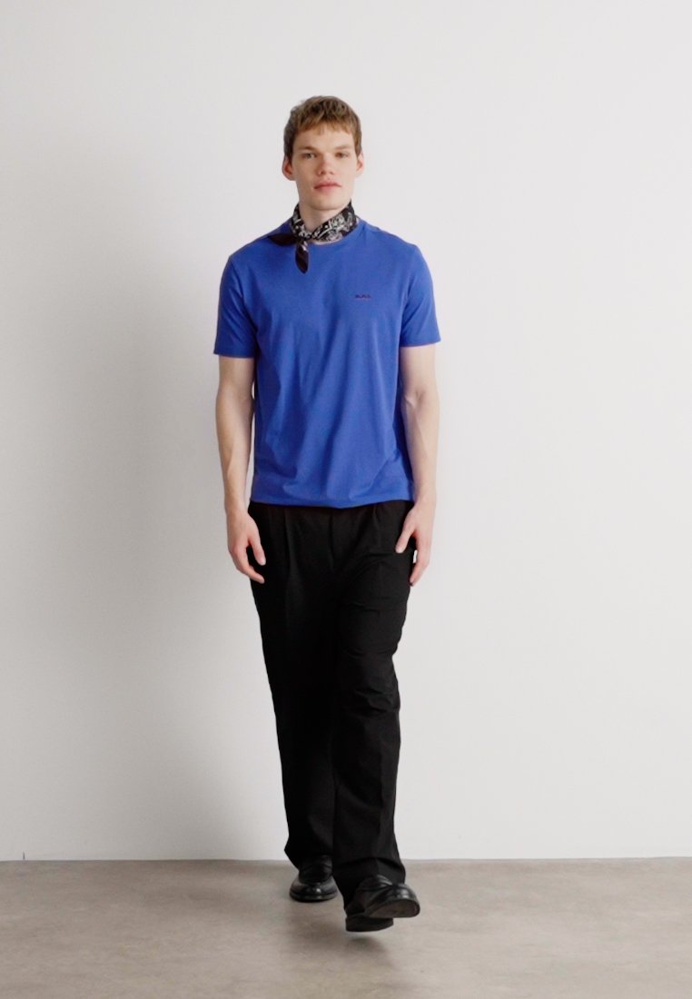 Un modèle portant un t-shirt bleu à manches courtes et un pantalon noir. Une écharpe à motifs est nouée autour du cou. Tissu lisse, coupe décontractée.