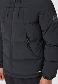 Melna puffer jaka ar rāvējslēdzēju priekšpusē, ar kapuci un sānu kabatām. Iezīmēta ar logo uz piedurknes un teksturētu audumu. Pieguļoša piegriezums.