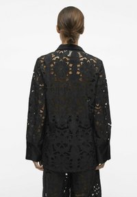 Veste en dentelle noire, avec des motifs floraux, des manches longues et un col en satin. Le matériau est transparent, révélant la peau en dessous.