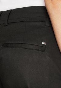 Black trousers with a smooth texture, featuring a back pocket and a small logo detail. The design includes a fitted waistband.