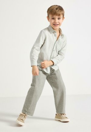 Hymyilevä nuori poika seisoo yhdellä jalalla eteenpäin, pukeutuneena vaaleaan raidalliseen paitaan, harmaisiin housuihin ja beigenvärisiin tennareihin yksinkertaisen valkoisen taustan edessä.