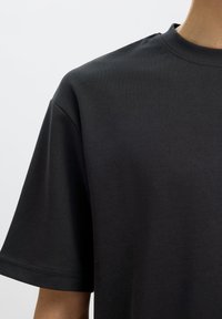 T-shirt noir en coton à manches courtes porté par une personne, montrant le haut de la poitrine et l'épaule gauche avec un col rond.