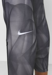 Legginsy Nike w ciemnoszarym kolorze z subtelnym abstrakcyjnym wzorem, z białym logo, detalami w postaci zamka oraz gładkim, elastycznym materiałem.