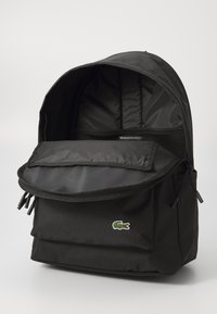 Sac à dos noir en tissu durable. Comprend une poche à rabat à l'avant, un intérieur en maille et un petit logo de crocodile brodé.