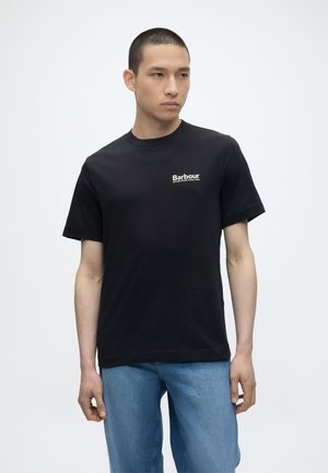 Νεαρός άνδρας που φοράει μαύρο t-shirt με το λογότυπο Barbour και ανοιχτόχρωμα τζιν, στέκεται μπροστά σε ένα απλό λευκό φόντο.