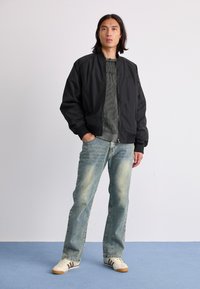 Veste bomber noire avec des poignets côtelés, pull gris texturé, jeans bleu délavé et baskets blanches avec des rayures brunes. Fond uni.