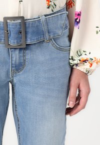 Light blue denim jeans with a wide, textured belt featuring a rectangular metallic buckle. Floral patterned long-sleeved blouse visible.