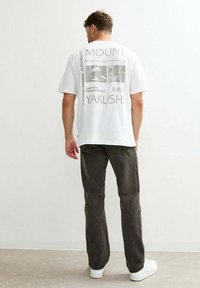Una persona sta in piedi con la schiena rivolta verso lo spettatore, indossando una maglietta bianca con grafiche e testo riguardanti il Monte Yakushi, abbinata a jeans scuri.
