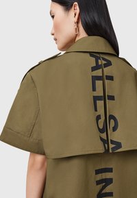 Oliwkowa, krótkozbrodowa koszula-kurtka z rozcięciem z tyłu, z dużym czarnym napisem "ALLSAIN." Gładki materiał z minimalnymi detalami.