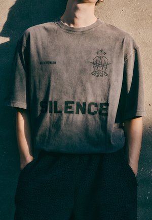 Personne portant un t-shirt gris foncé délavé avec le mot "SILENCE" imprimé en grand sur le devant et les mains dans les poches d’un pantalon noir texturé.