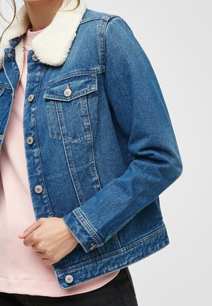 Personne portant une veste en jean bleue avec un col sherpa blanc sur une chemise rose, ajustant l'ourlet de la veste de la main.
