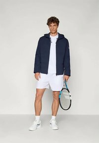 Jūras spēku jakas ar kapuci pār baltu T-kreklu, kombinētas ar baltām šortiem. Modelis tur tenisista raketi un valkā baltas sporta kurpes.