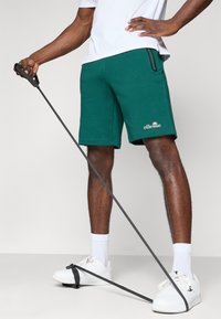 Grønne atletiske shorts med sidelommer og et logo. Båret med hvide sneakers og sokker. En modstandsbånd holdt i den ene hånd.