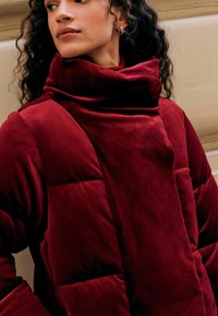 Burgundy puffer jacket with a high collar and quilted sections. Soft, velvety texture with angular seams, highlighting a modern design.