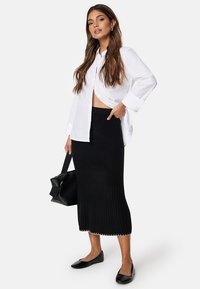 Zwarte ribgebreide rok met een uitlopende zoom, gecombineerd met een witte blouse met knopen. Model draagt een zwarte tas en heeft zwarte instappers aan.