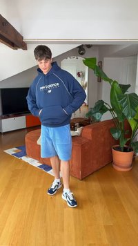 Young man wearing navy New Balance hoodie, blue shorts, and sneakers standing in a modern living room next to an orange couch and large potted plant.