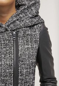 Schwarze und weiße Kapuzenjacke aus einem strukturierten Gewebe mit einem Zickzackmuster und schwarzen Lederärmel. Mit einem Frontreißverschluss.