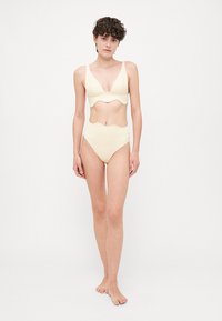 Bežs bikini komplekts, kas izgatavots no gludas auduma. Augšdaļai ir viļņota mala, bet apakšdaļas ir augstvērtīgas ar viļņotu dizainu.