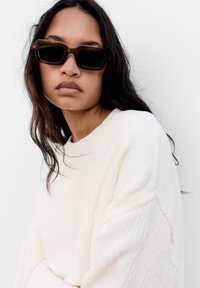 Barna téglalap alakú napszemüveg sötét lencsékkel. Egy modell krémszínű, texturált pulóvert visel, világos háttér előtt. Természetes, hullámos haj.